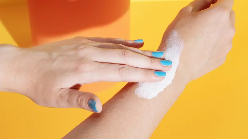 A woman's hand rubbing sunscreen on her arm against an orange background.