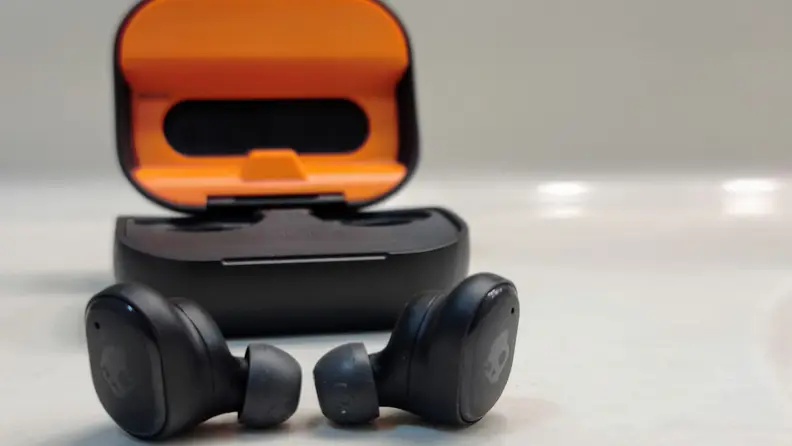 The all-black Skullcandy Grind Fuel sit in front of their orange-lined case on a white reflective glass table with the eartips pointed in.