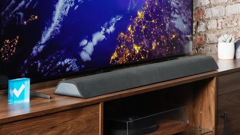 Angled shot of the Vizio M-Series All-in-One M213AD-K8 Dolby Atmos Soundbar resting on an entertainment center in front of a TV.