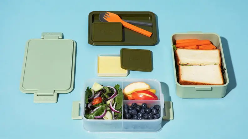 A green Bento box with an assortment of food including a sandwich, fruit, and vegetables.