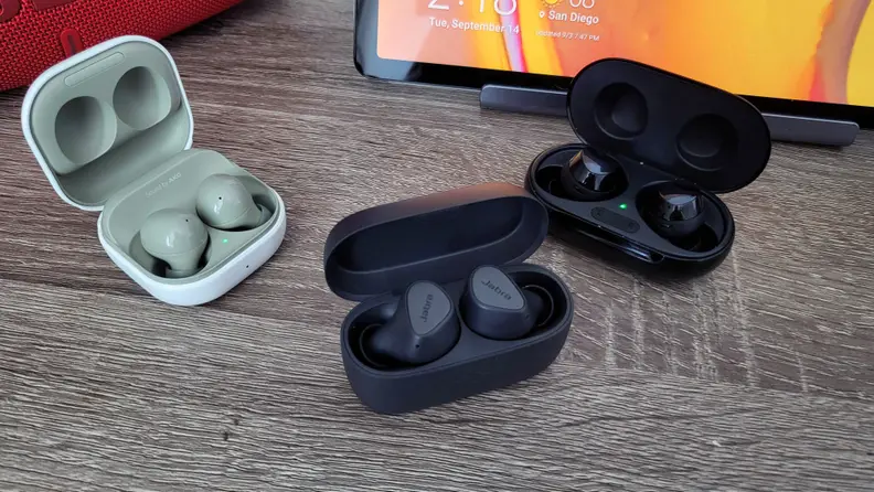 Jabra's all-black, matte earbuds sit in their open case between the slick and glossier Galaxy Buds 2 and Galaxy Buds Plus on a wooden surface.