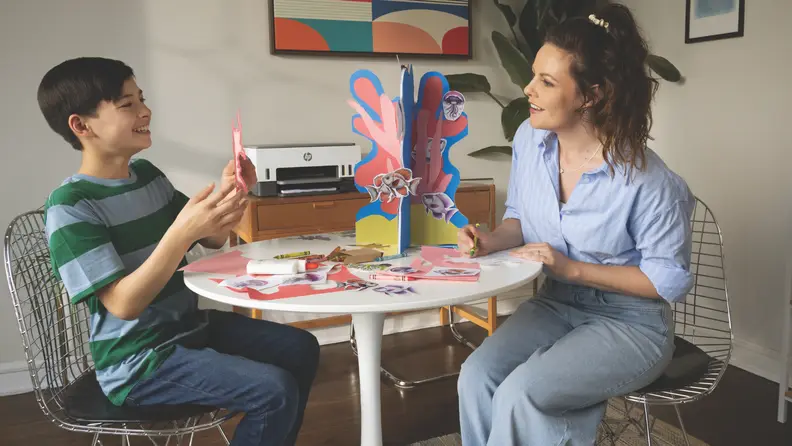 A mom and child doing arts and crafts at a table with an HP printer in the background.