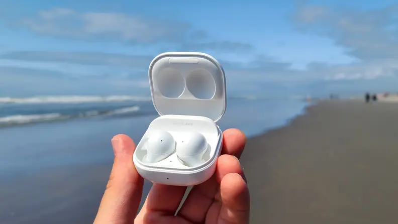 Samsung Galaxy Buds 2 are held in their white case with the micro USB showing against a sunny beach and a blue ocean.