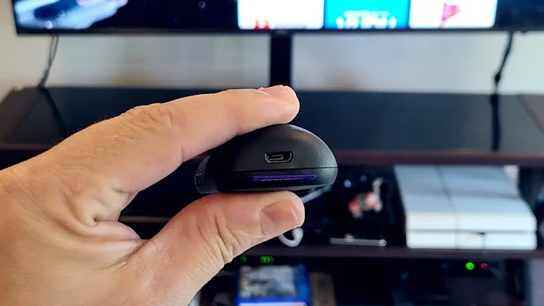A hand holds the black, rounded remote in front of a TV console with a microUSB port showing.