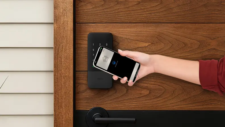 A person holds a smartphone near a keypad-style smart lock mounted on a wooden door, demonstrating contactless unlocking.