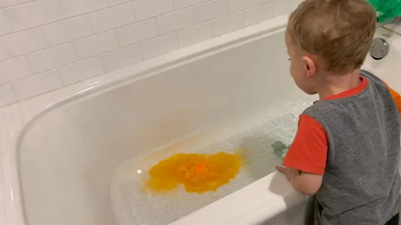 A small child inspects a bathtub as the water turns orange, thanks to the dye from a bath bomb.