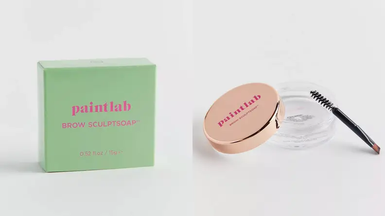 On the left: A small, light green box with pink lettering on it. On the right: A clear jar with clear brow gel in it uncapped with its gold lid and spoolie brush leaning on the jar.