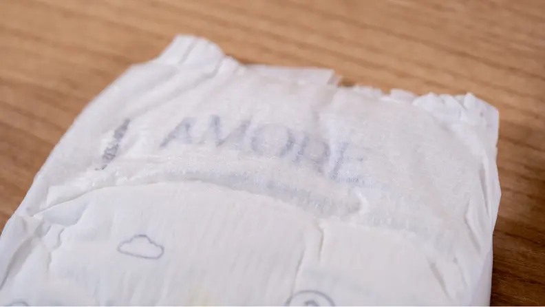 Top half of Pampers Amore diaper shown with "Amore" text, placed on top of a wooden surface.