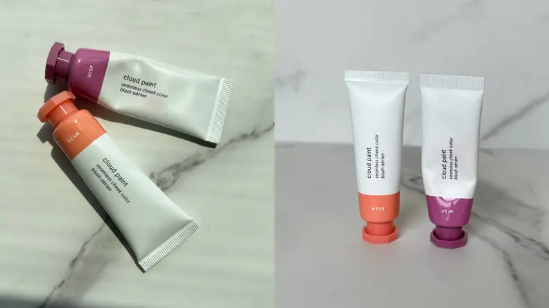 Two tubes of Glossier Cloud Paint in orange and purple shades against a marble-textured background.