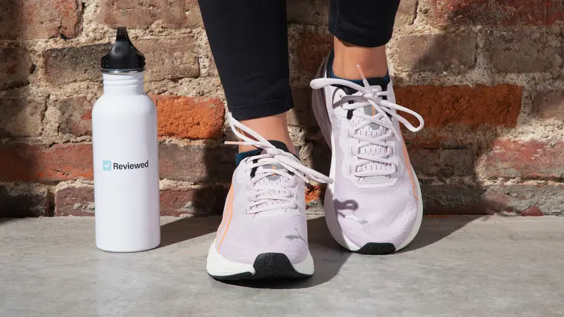 A person leans against a brick wall wearing the Puma Run XX Nitro running shoes standing next to a white water bottle.