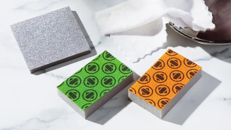 Product shot of the green and orange Skura Style sponge next to microfiber cloths on marble countertop.