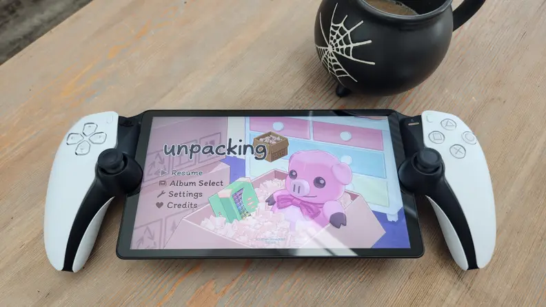 A white handheld gaming device, the playstation portal, on top of a light brown wooden surface with a black mug engraved with a spiderweb above it.