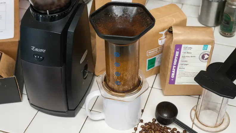 Assorted coffee gadgets arranged on a white surface, including a coffee mug, an Aeropress coffee maker, a coffee bean grinder, a measuring spoon, and coffee bean bags.