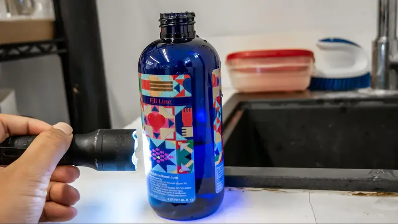A person holding a flashlight and shining it into a dark blue bottle of Truly Free Space Freshener with a colorful graphic label.