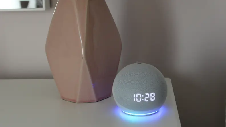 An Amazon Echo Dot sits on a nightstand next to a bedside lamp.