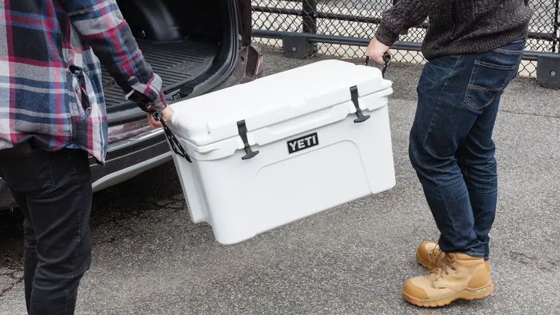 Two people carrying a Yeti cooler to a car