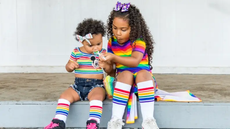 Two little girls sitting together outdoors sharing a snow cone while wearing rainbow attire.