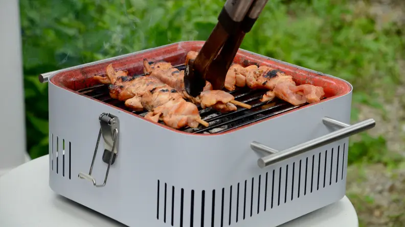 A person is using a pair of tongs to grab a chicken skewer, which is being cooked on an Everdure Cube grill in gray color.