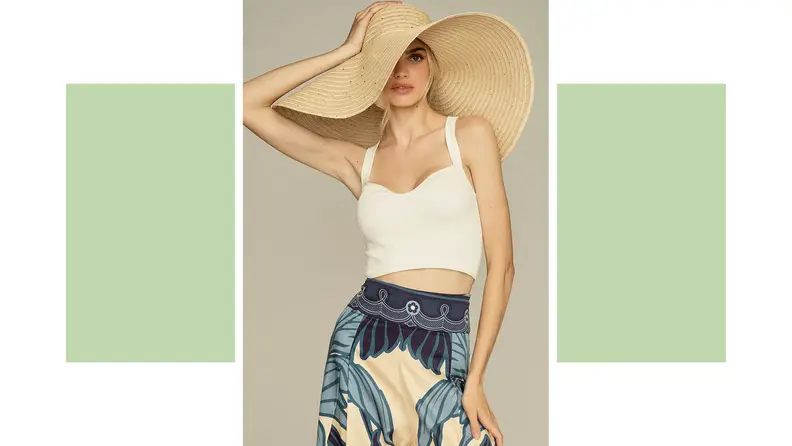 A woman wears an oversized straw sunhat with a white tank and denim skirt.