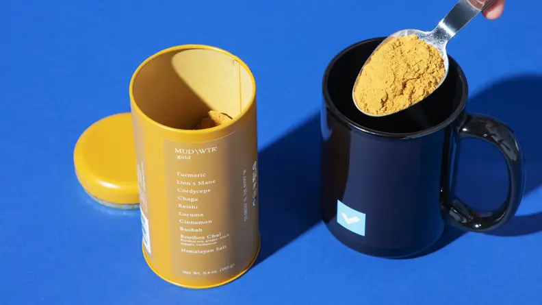 Mud/Wtr product containers, in the color blue and yellow, with the lid off and a scoop of powder being removed from the container.