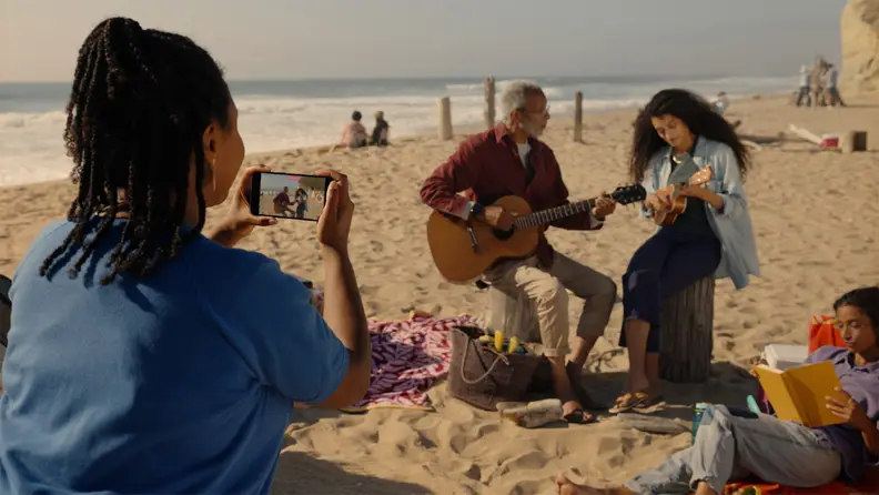 A person using their iPhone to record two people playing instruments on the beach on a sunny day.