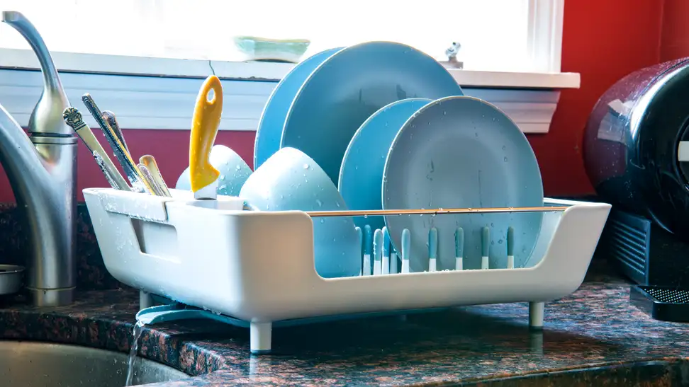 A dish rack filled with blue plates, cups and utensils sits next to a kitchen sink.