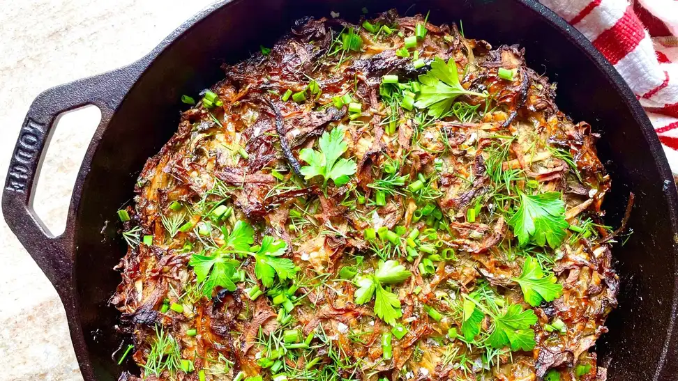 A close-up overhead photo of crispy potato kugel in a Lodge cast iron skillet, garnished with fresh parsley and dill.