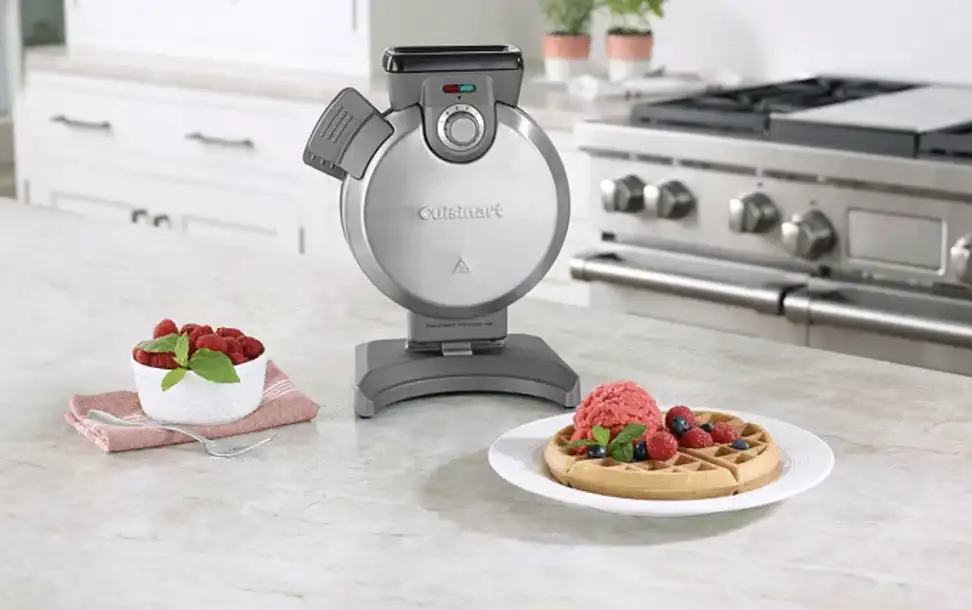 A vertical, Cuisinart waffle maker, sitting on a countertop next to a freshly made waffle with strawberry topping.
