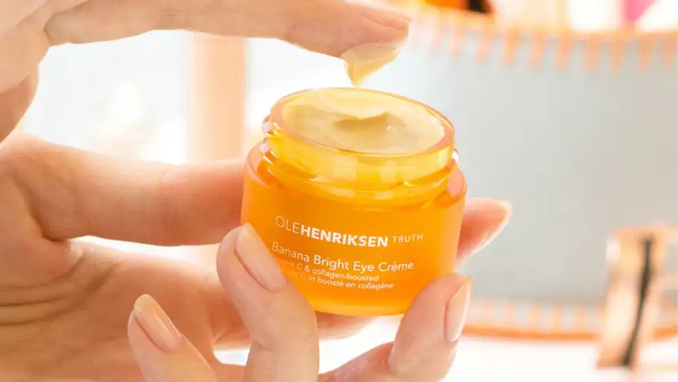 A hand holding the orange-colored OleHenriksen Banana Bright Eye Crème jar. The jar is open to reveal a light orange cream and an index finger is dipping into the pot to pick up cream.