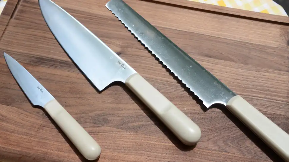 On a walnut wood cutting board by Our Place, a set of knives are on display. From the left, there's a paring knife, a chef's knife, and a serrated slicing knife.