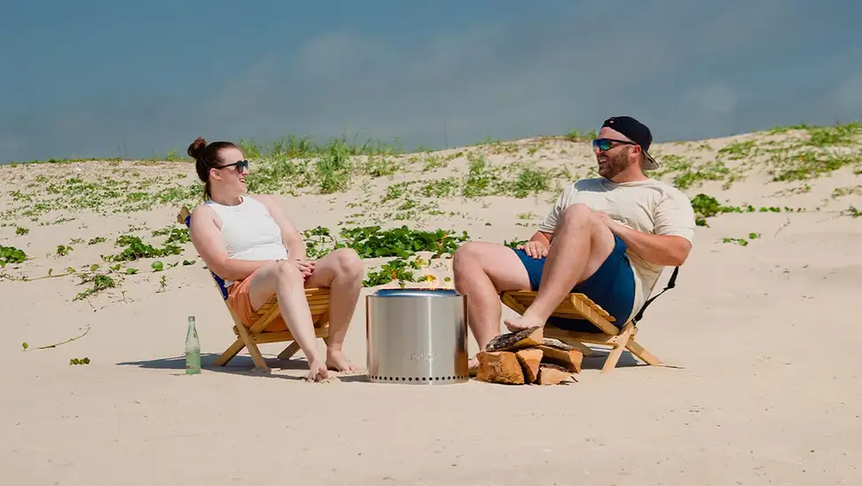 A couple sit around an outdoor fire pit on a beach.