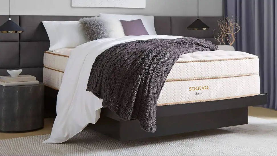 Saatva mattress sitting in bedroom with blanket and pillows sitting on top.