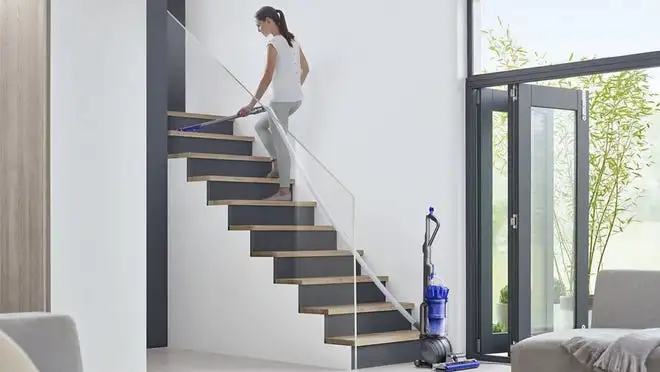 A person walks up stairs inside a home. A Dyson vacuum cleaner sits upright on the floor.