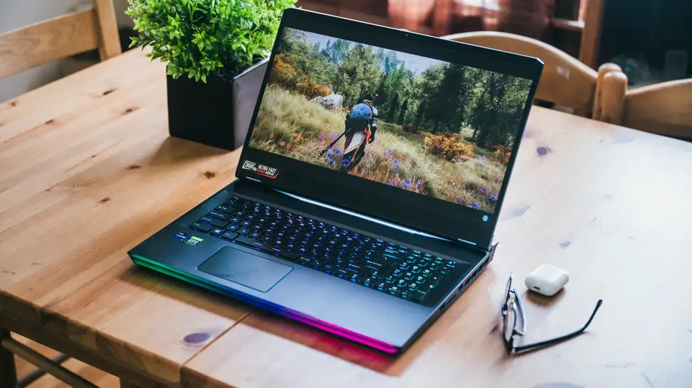 A laptop with a colorful base sits on a wooden desk in fresh sunlight next to a plant.