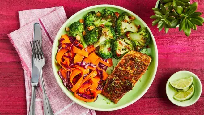 Plate filled with carrots, broccoli and salmon next to small plant, cutlery and limes on top of table.