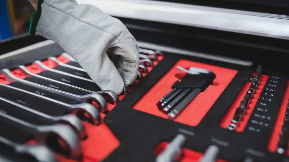 A work-gloved hand reaches into an open toolkit to grab a ratchet. The ratchets are organized in order of size. The tool kit is red with black detailing.