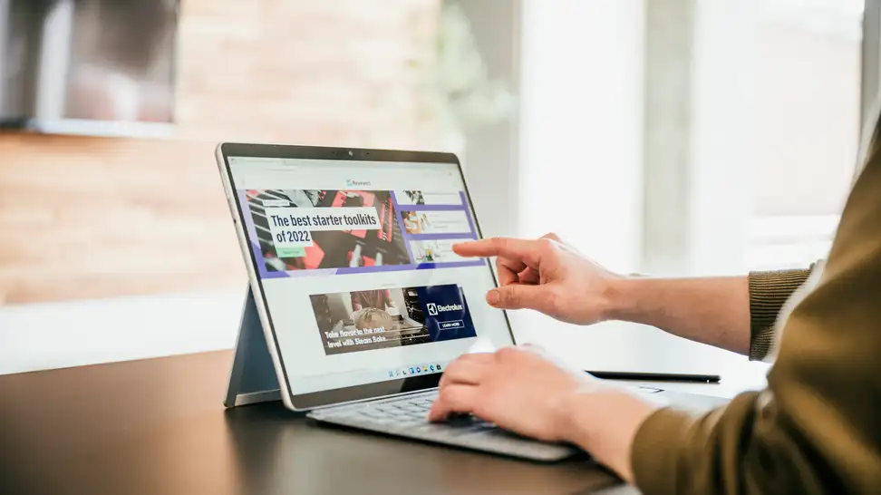 Microsoft's Surface Pro 8, with the kickstand extended, showing Reviewed.com's homepage, with a hand touching the display.