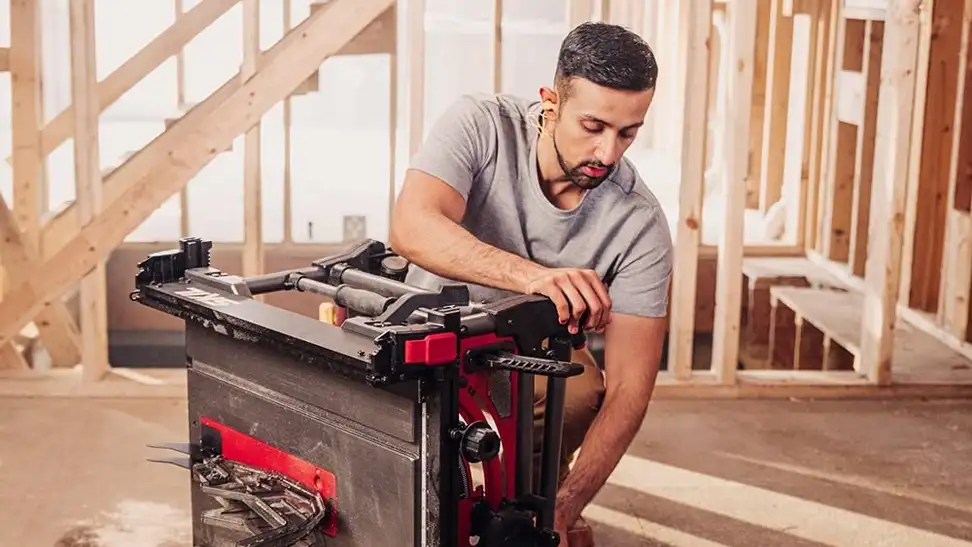 A model in a gray T-shirt demonstrates a Skil table saw at a construction site.