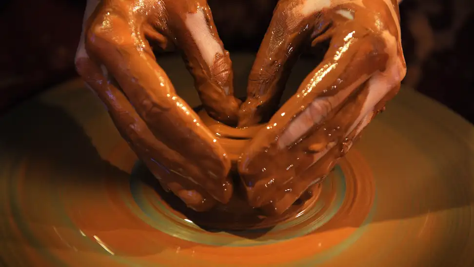 Hands mold wet, brown clay on a spinning pottery wheel.