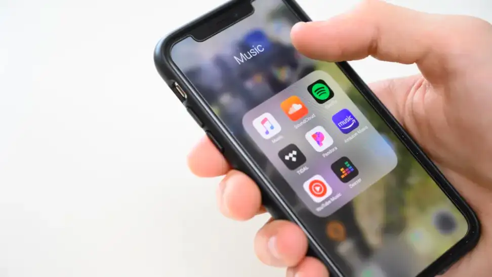 A hand holding an iPhone which displays the music folder and several popular music streaming apps.