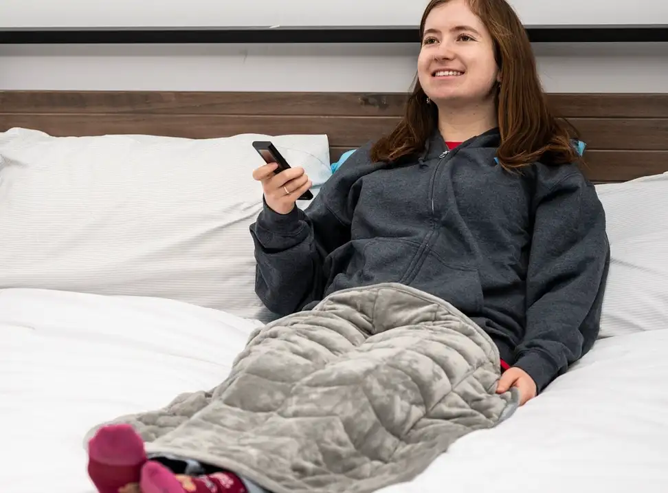 Woman laying on bed holding remote with grey blanket over her legs