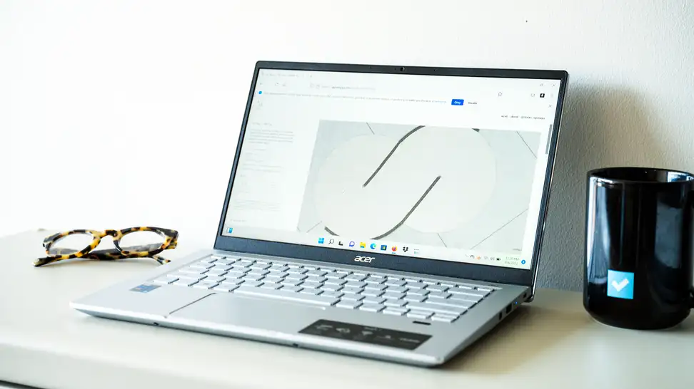 An open and powered on silver laptop sitting on a white surface with a black mug on the right and a pair of glasses on the left