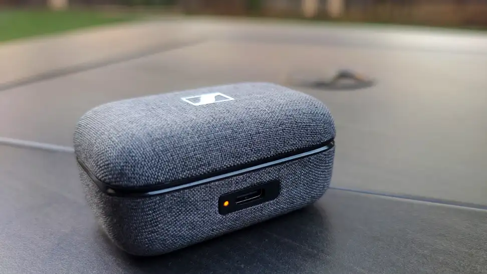 A gray fabric earbuds case sits on a black table with an orange LED at the front.