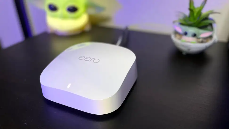 A white internet router box on top of a black table with two brightly colored objects in the background.