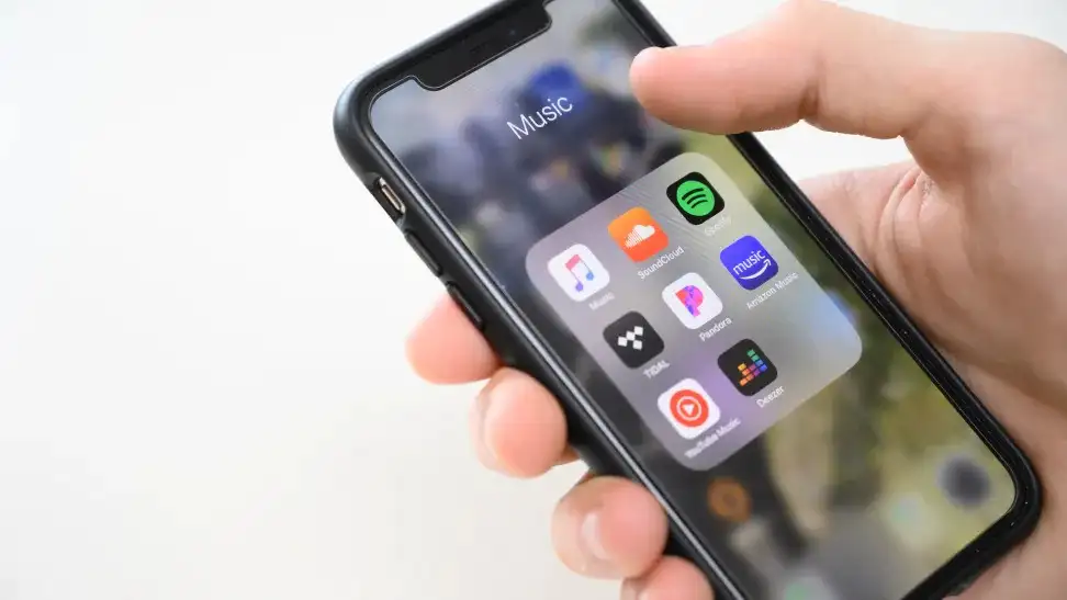 An iPhone X being held, with a folder of music apps shown on-screen. The apps include Apple Music, Soundcloud, Spotify, Tidal, and Pandora.
