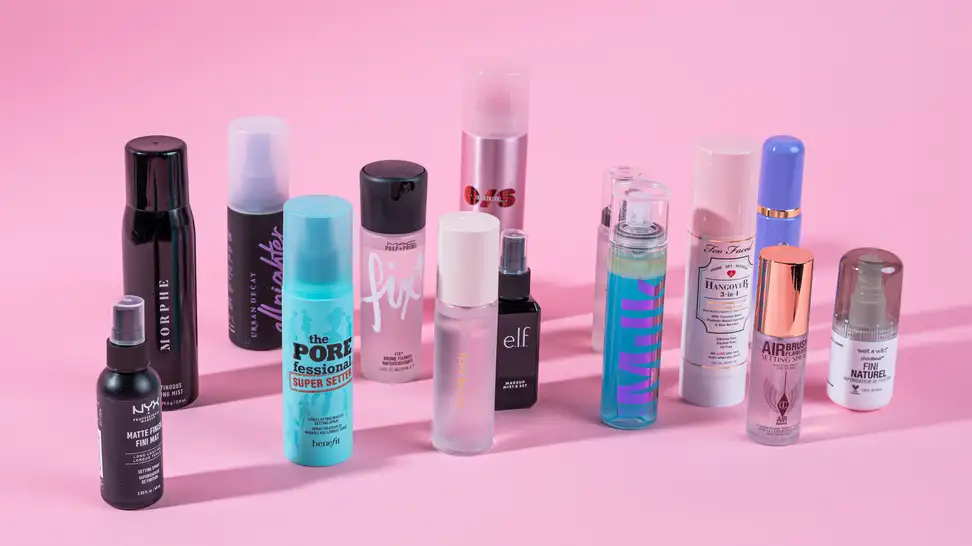 A bunch of makeup setting sprays standing on a pink background.