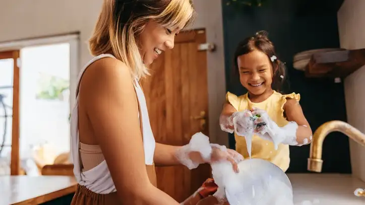 An adult and child washing dishes together and smiling