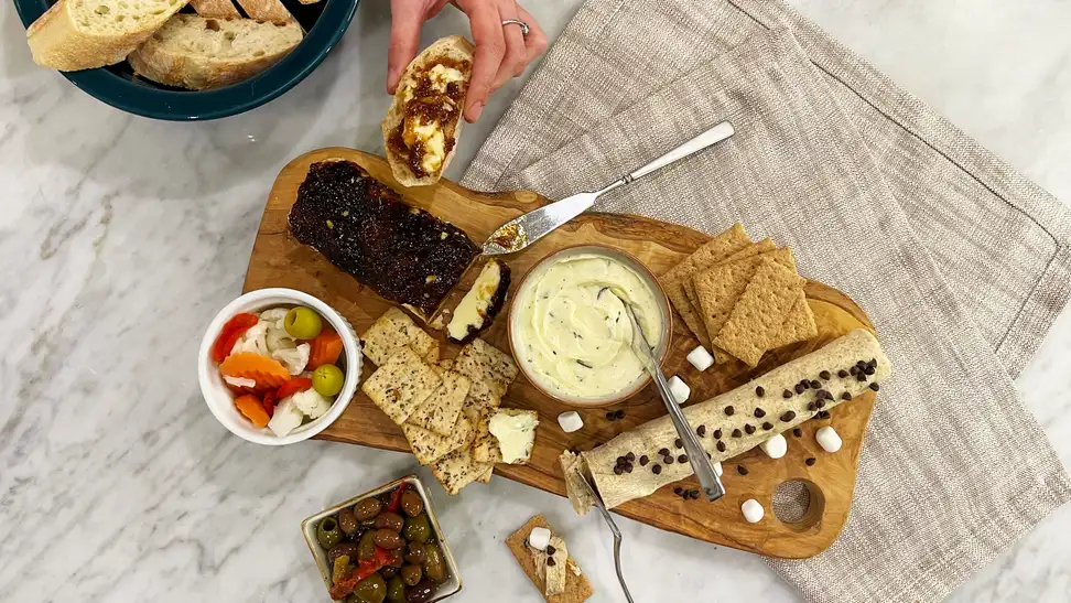 A hand holding a piece of bread with butter beside a butter board with accompaniments