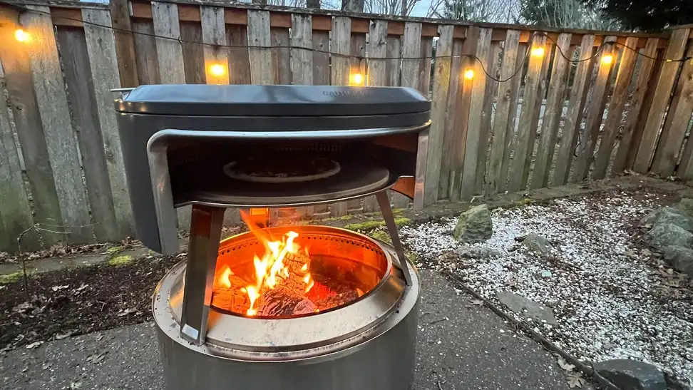 Solo Stove Pi Fire pizza oven with open flame outdoors in front of wooden fence with string lights.