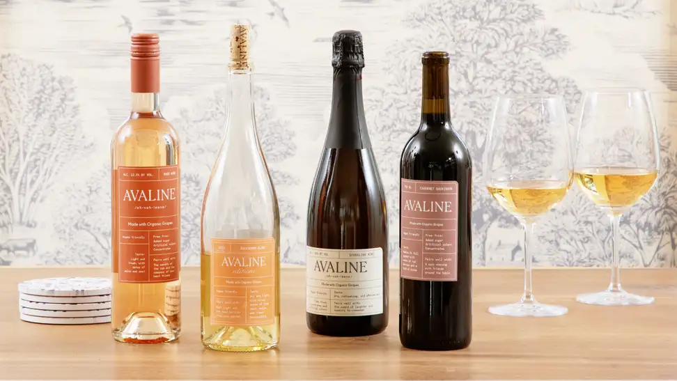 Two white wine and two red wine bottles of Avaline wine and two glasses of white wine on a wood table against a toile background.